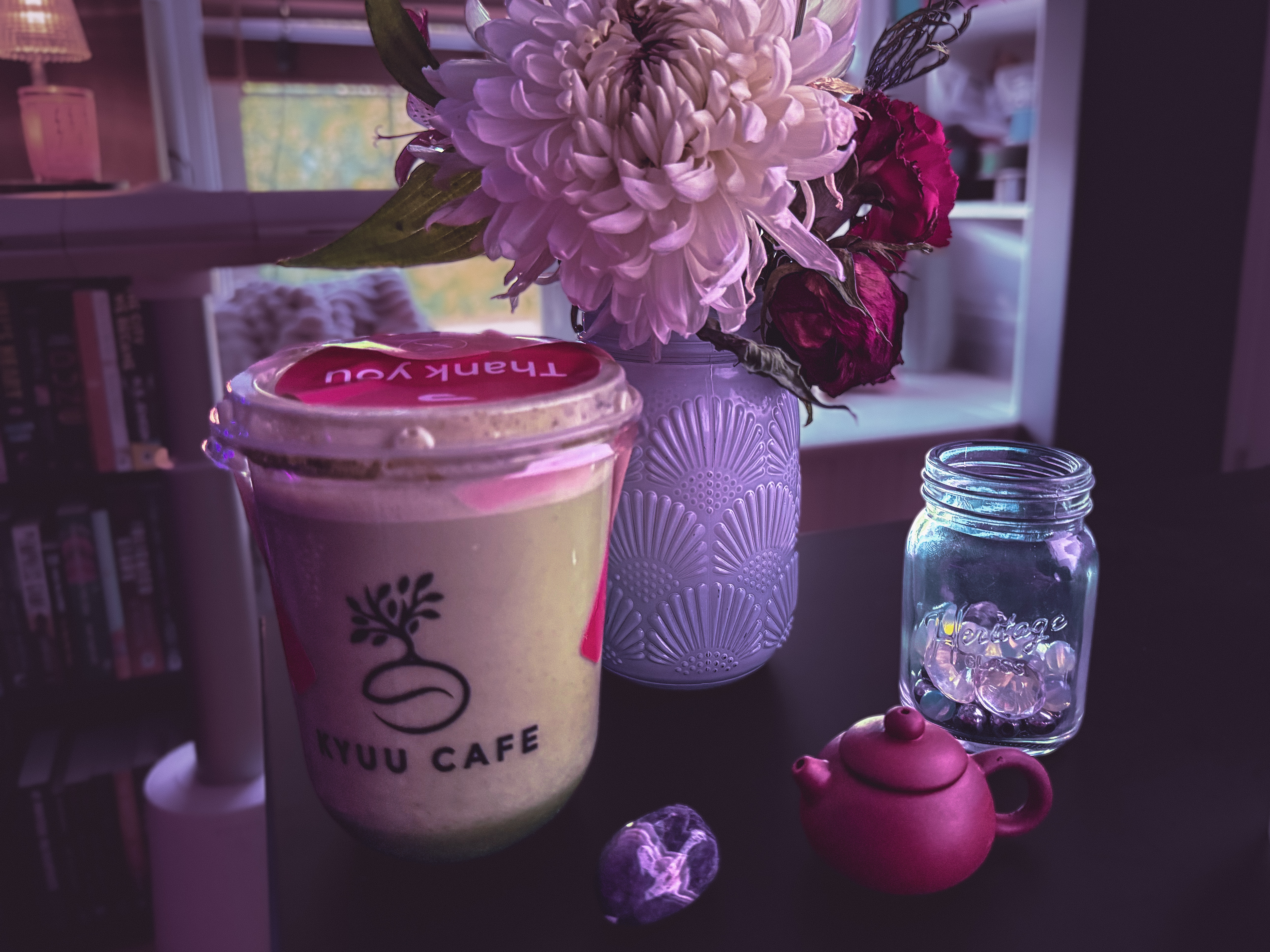 Aven's writing desk, with the following: an iced matcha from Kyuu Cafe, a white jar with white and red flowers, a tiny ceramic teapot, and a jar of moonstone beads.