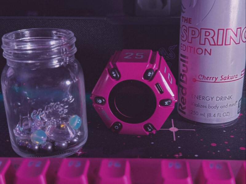 Aven's Desk A desk setup, with a magenta keyboard, Sakura Red Bull, pink hexagonal pomodoro timer, and Aven's progress jar filled with silver and moonstone beads.